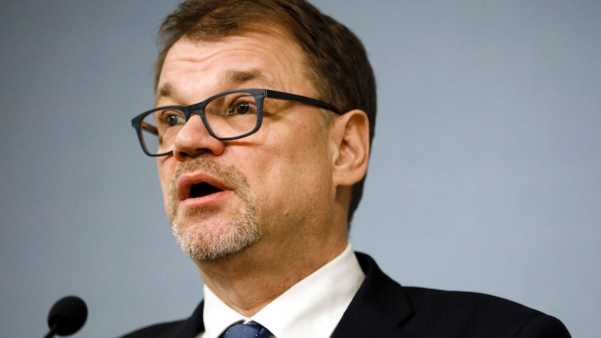 A Caucasian man in a black suit and blue tie with black-rimmed glasses speaks in front of a teal background.