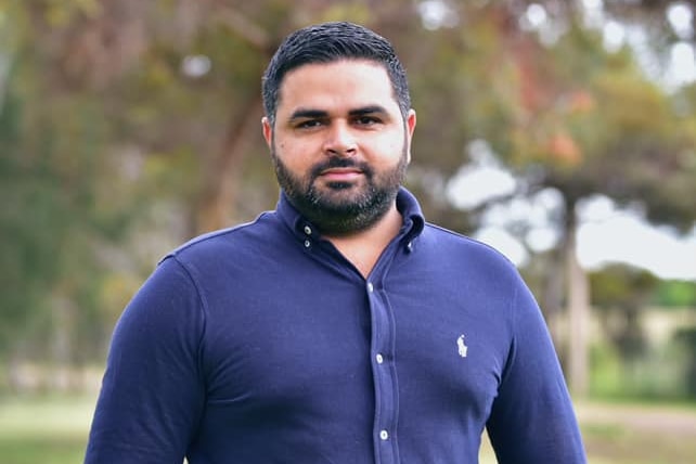 A man with black hair and a beard standing up wearing a blue shirt