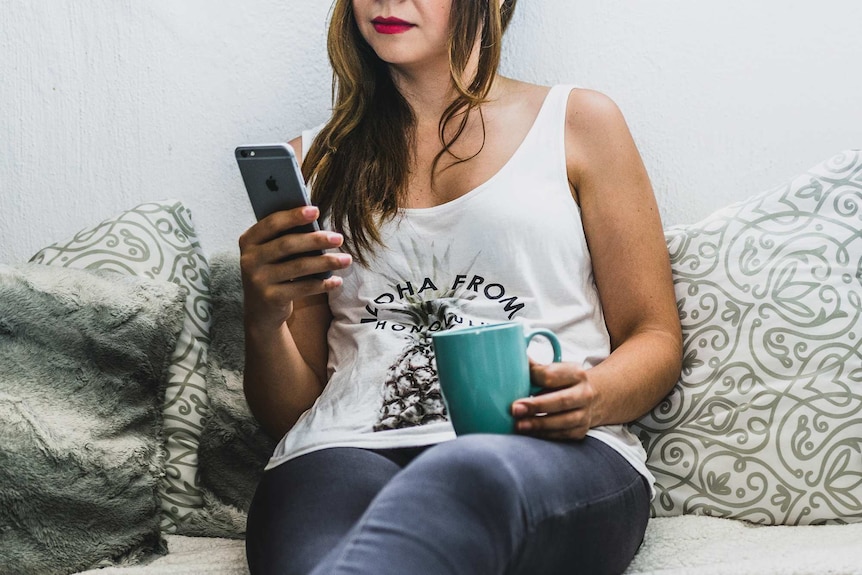 Woman sitting in bed looking at her smartphone, with a laptop, to depict the use technology to procrastinate from sleep.