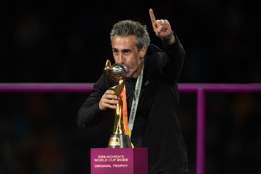 A man wearing a black jacket kisses a gold trophy and holds his finger in the air