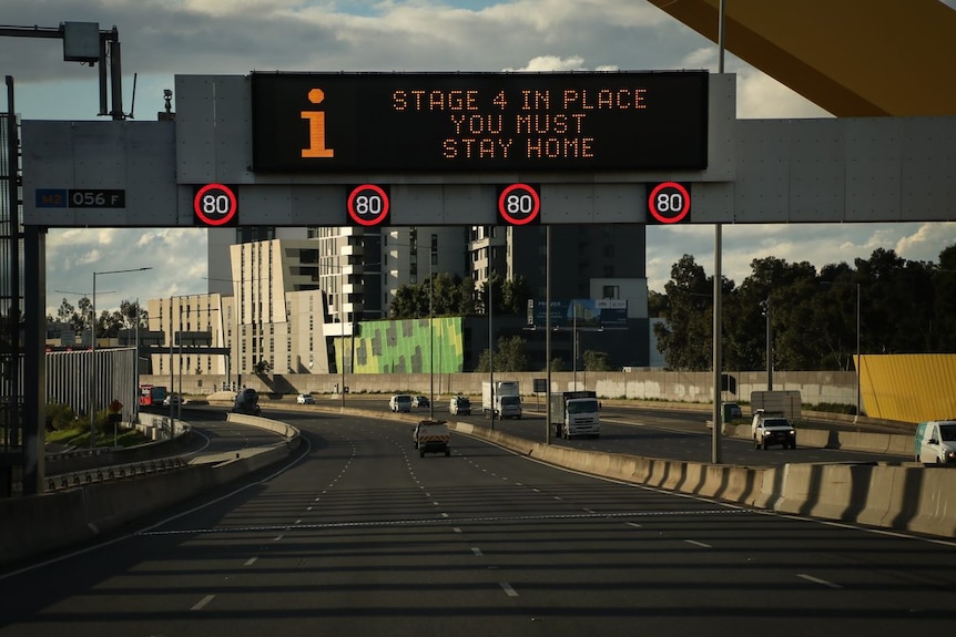 A toll road with minimal traffic and a gantry sign saying 'stage 4 in place you must stay home'.