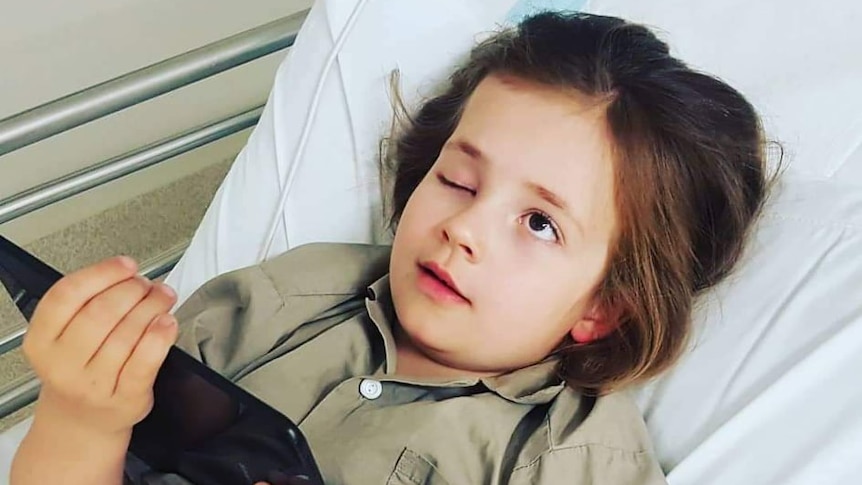 A young girl with one eye lid permanently closed on a hospital bed with an iPad in her hands