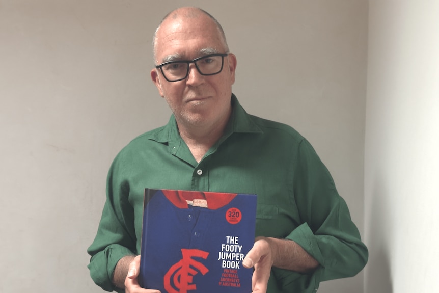 A middle-aged man, balding, glasses, holding his red and blue football jumper book wearing a green linen shirt. He is smirking.