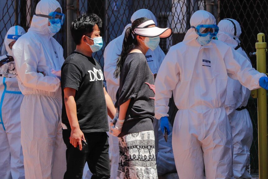 A number of people in white protective gear usher two people in plain clothes and masks to a fenced area