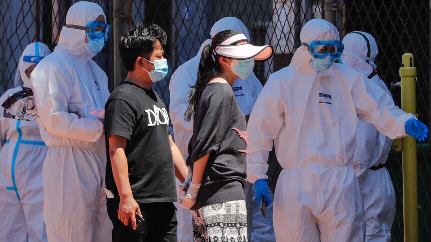 A number of people in white protective gear usher two people in plain clothes and masks to a fenced area