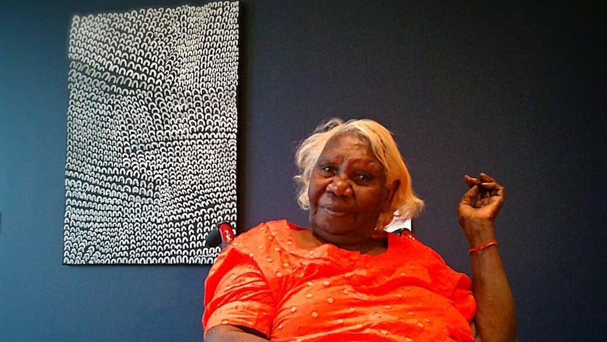 an Indigenous woman in an orange dress smiles as she sits in front of a black and white painting 