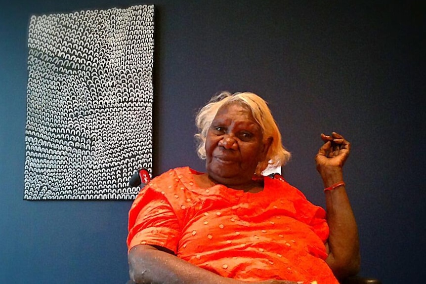 an Indigenous woman in an orange dress smiles as she sits in front of a black and white painting 