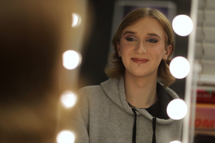 A teenage girl wearing eye-liner and makeup smiles, looking at herself in a mirror. The mirror's frame has lightbulbs.
