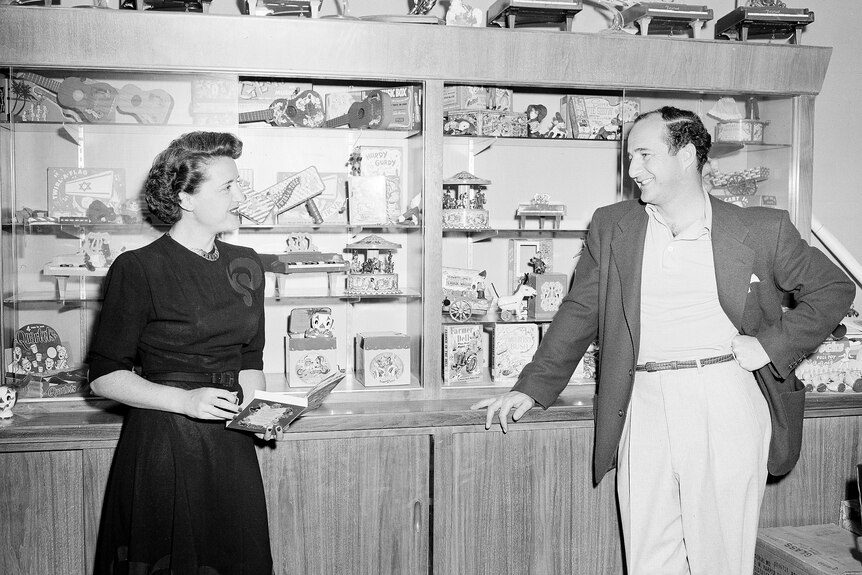 A black and white photo of a woman dressed in a black dress looking over at a man dressed in a suit near a display of toys.