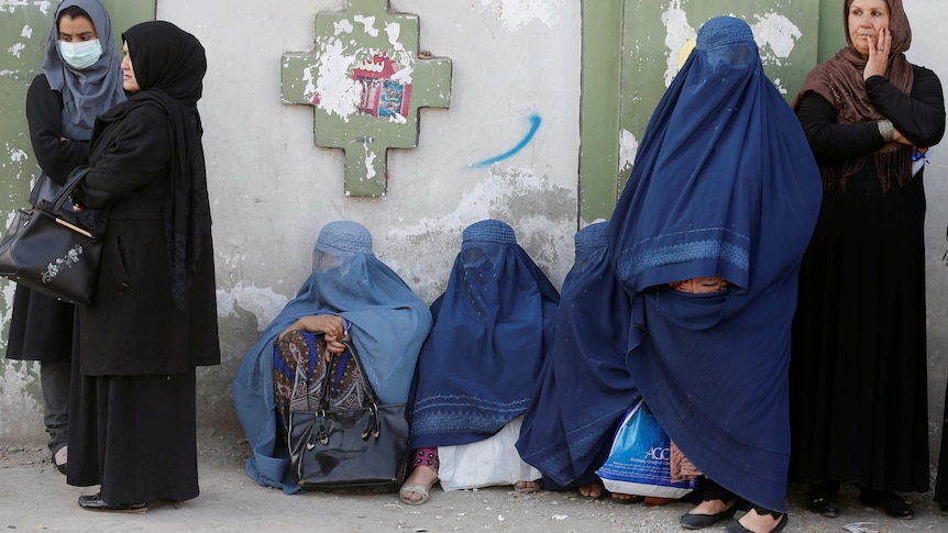 A group of women stand against a wall with some wearing burqa coverings and others in headscarves.