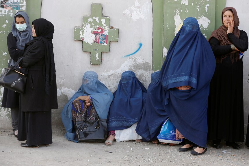 A group of women stand against a wall with some wearing burqa coverings and others in headscarves.
