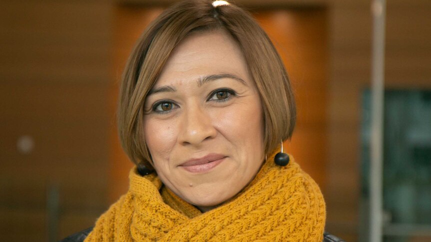 A woman in her early 40s smiles into the camera wearing her hair in a short bob, a cable knit yellow scarf around her neck.