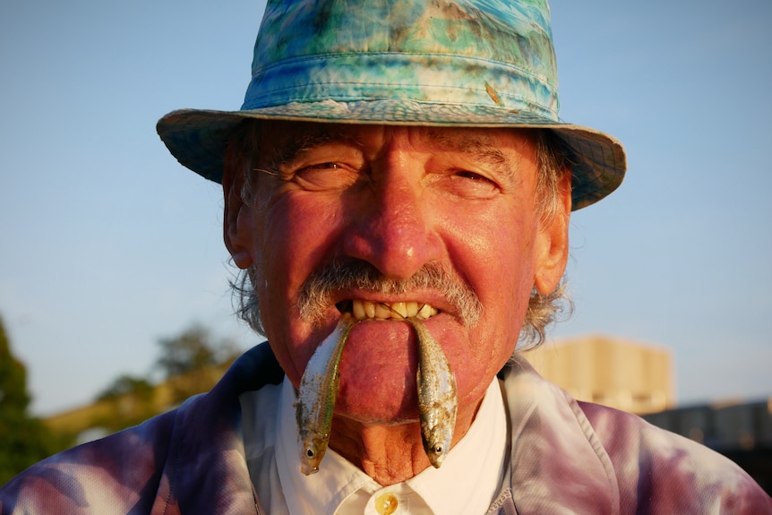 A man in a hat smiles with two small fish hanging from his teeth.