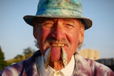 A man in a hat smiles with two small fish hanging from his teeth.