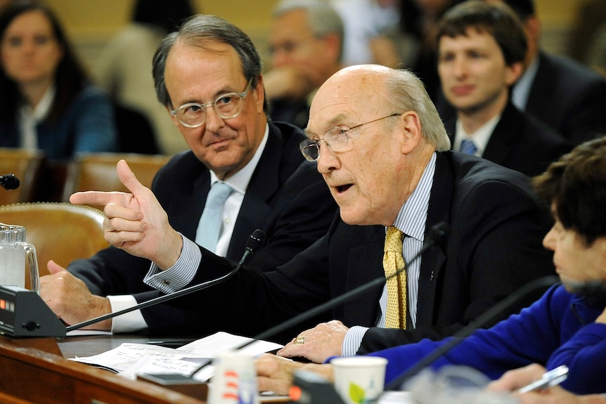 an elderley man speaks into a desk microphone pointing his finger forwards