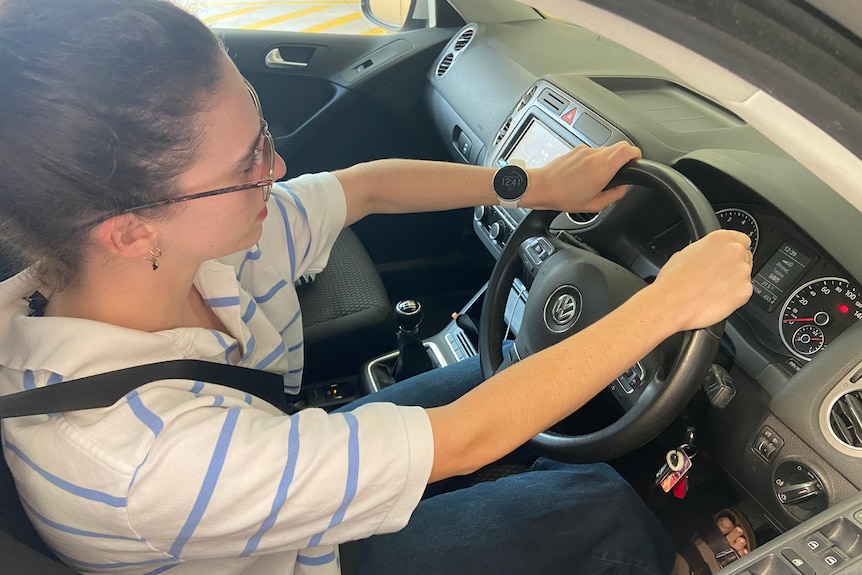 An overhead shot of a young woman wearing a white t-shirt, blue stripes, hands on steering wheel, a smart watch on her wrist.