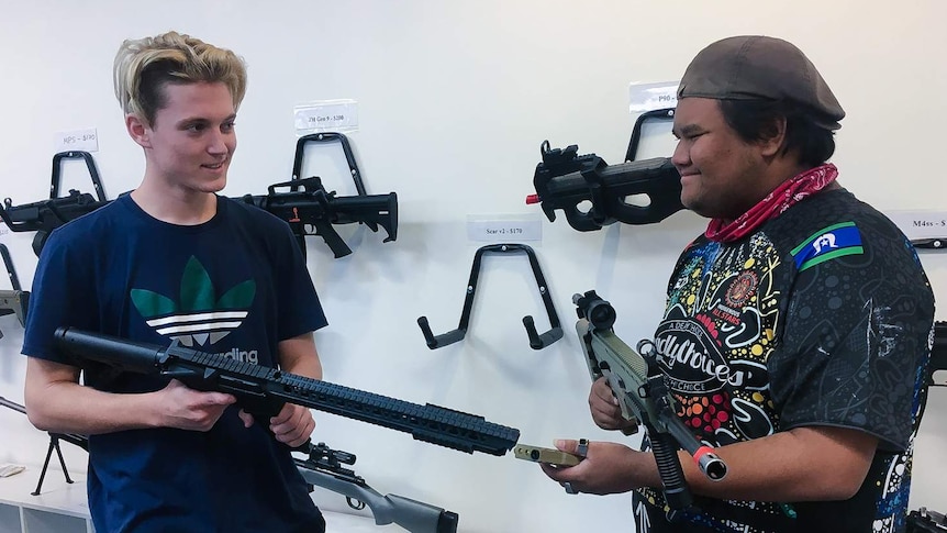 Two young men hold replica firearms while smiling at each other. Gel blaster 'gun's hang on wall in background.