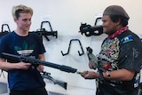 Two young men hold replica firearms while smiling at each other. Gel blaster 'gun's hang on wall in background.