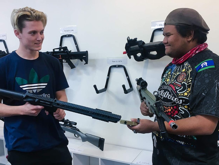 Two young men hold replica firearms while smiling at each other. Gel blaster 'gun's hang on wall in background.