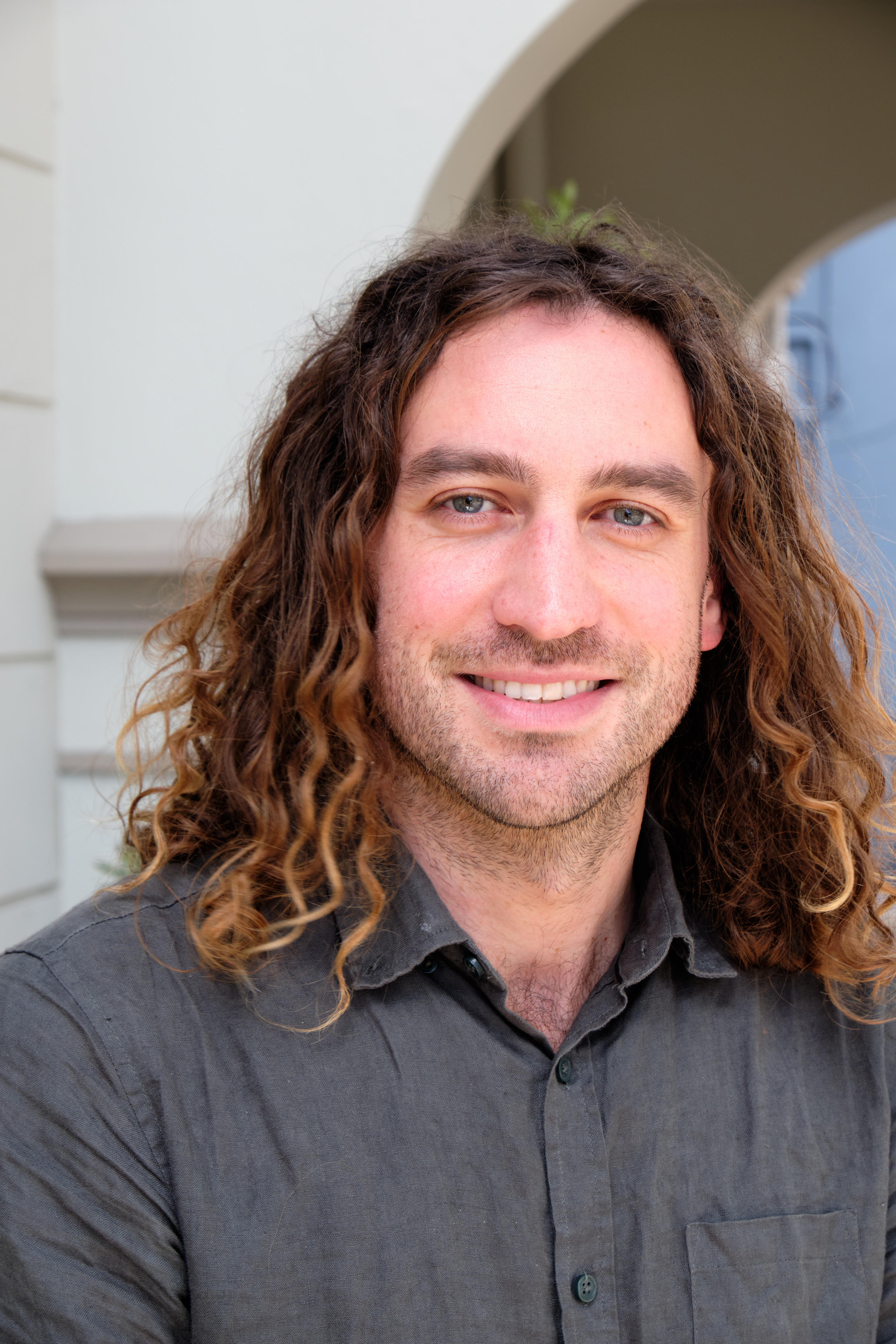 Um homem com cabelos ondulados na altura dos ombros sorri para a câmera.