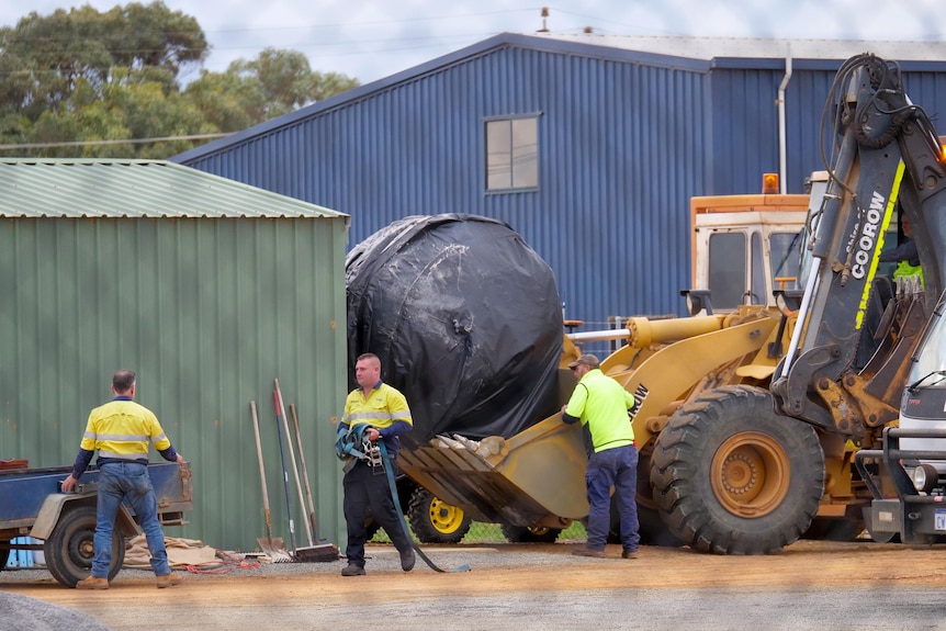Workmen around a front-end loader depositing a large object wrapped in plastic into a shed.