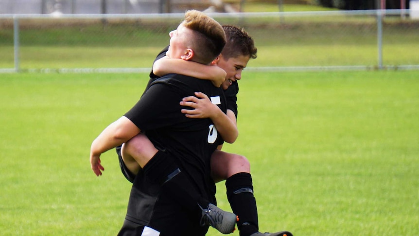 A boy kitted up in soccer gear on a soccer pitch jumps on another teammate in celebration.