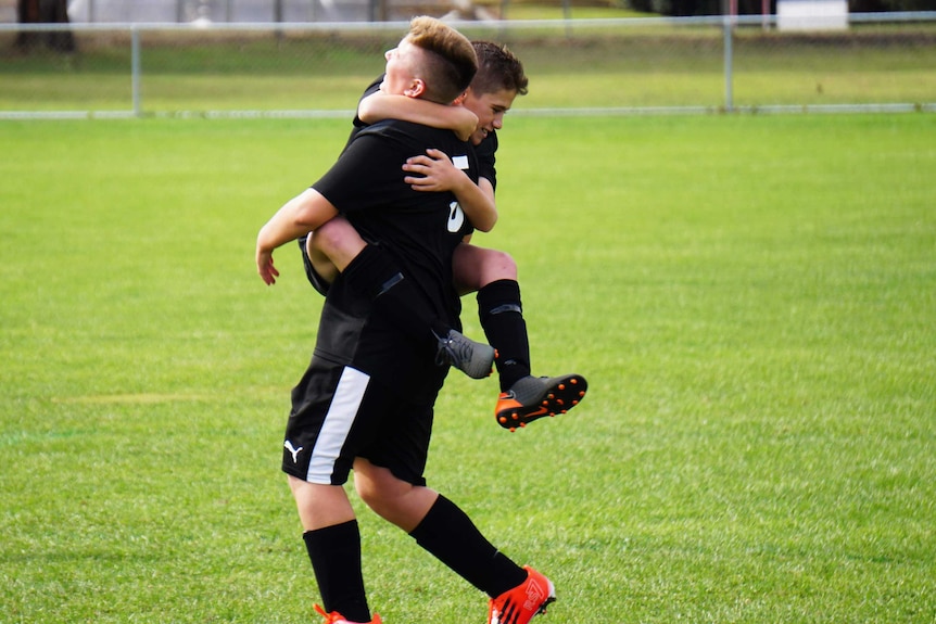 A boy kitted up in soccer gear on a soccer pitch jumps on another teammate in celebration.