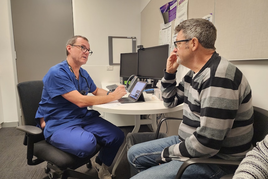 A bespectacled man in scrubs sits in an office, speaking to another man.