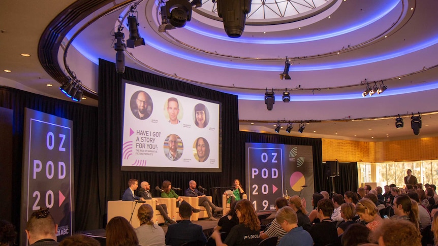 Wide shot of panel and audience listening.