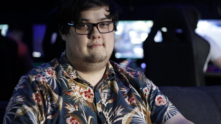 A young man with glasses and a button up floral shirt smiles with computers behind him.