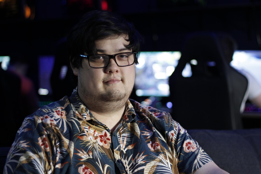 A young man with glasses and a button up floral shirt smiles with computers behind him.