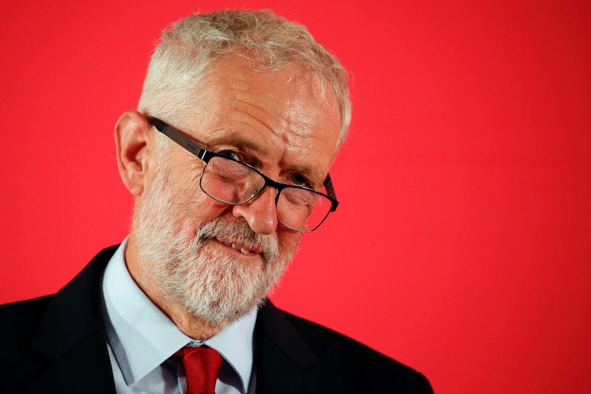 Jeremy Corbyn, standing against a red background, tilts his head.
