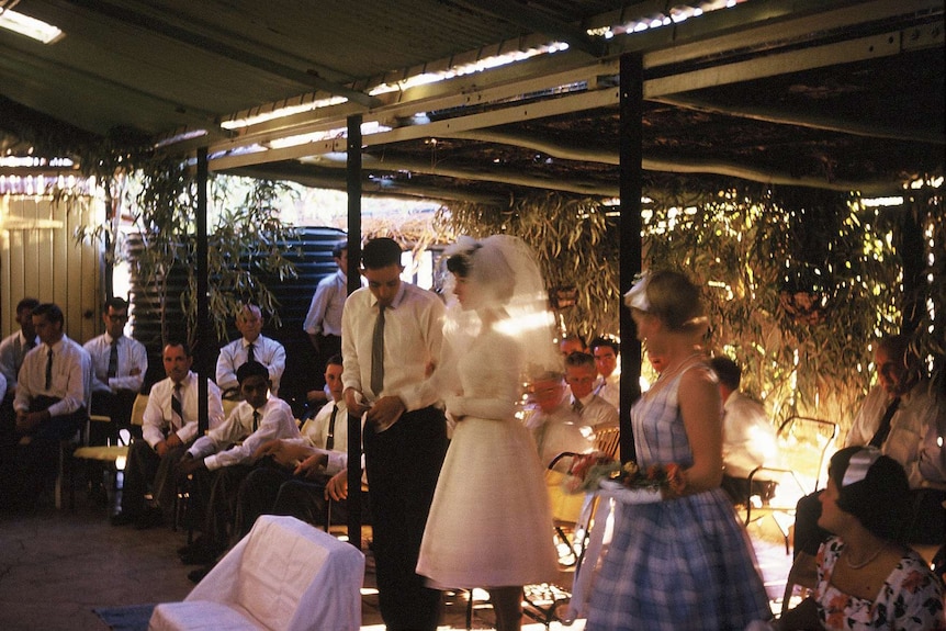a bride and groom surrounded by wedding guests standing in a shed