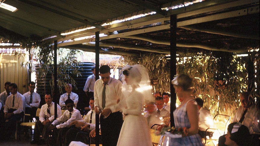 a bride and groom surrounded by wedding guests standing in a shed