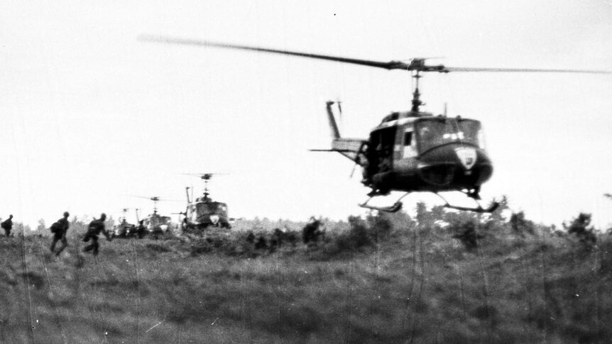 Helicopters hovering above a field with silhouettes of soldiers in the distance.