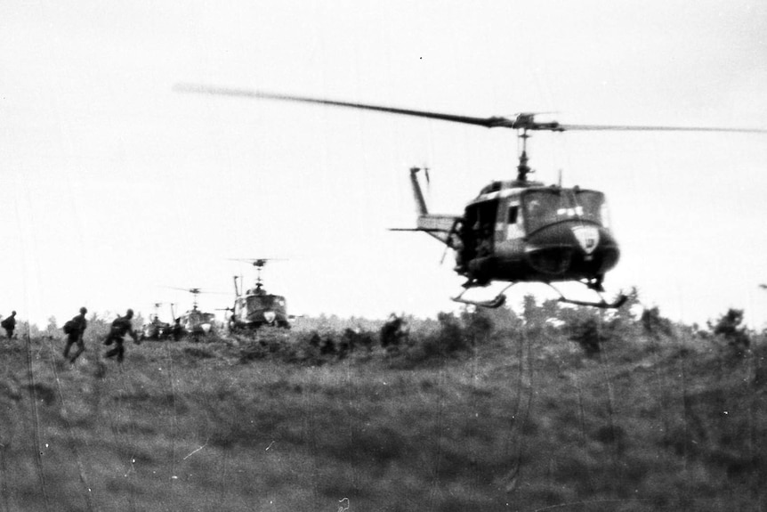 Helicopters hovering above a field with silhouettes of soldiers in the distance.