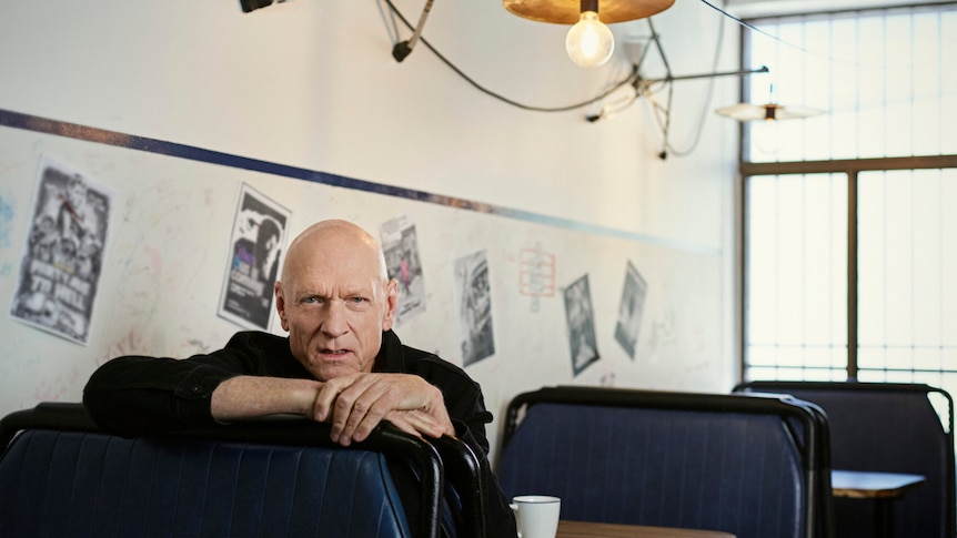 Peter sits in a diner-style cafe, looking over the back of a chair at the camera. He has a cup of coffee and is wearing black.