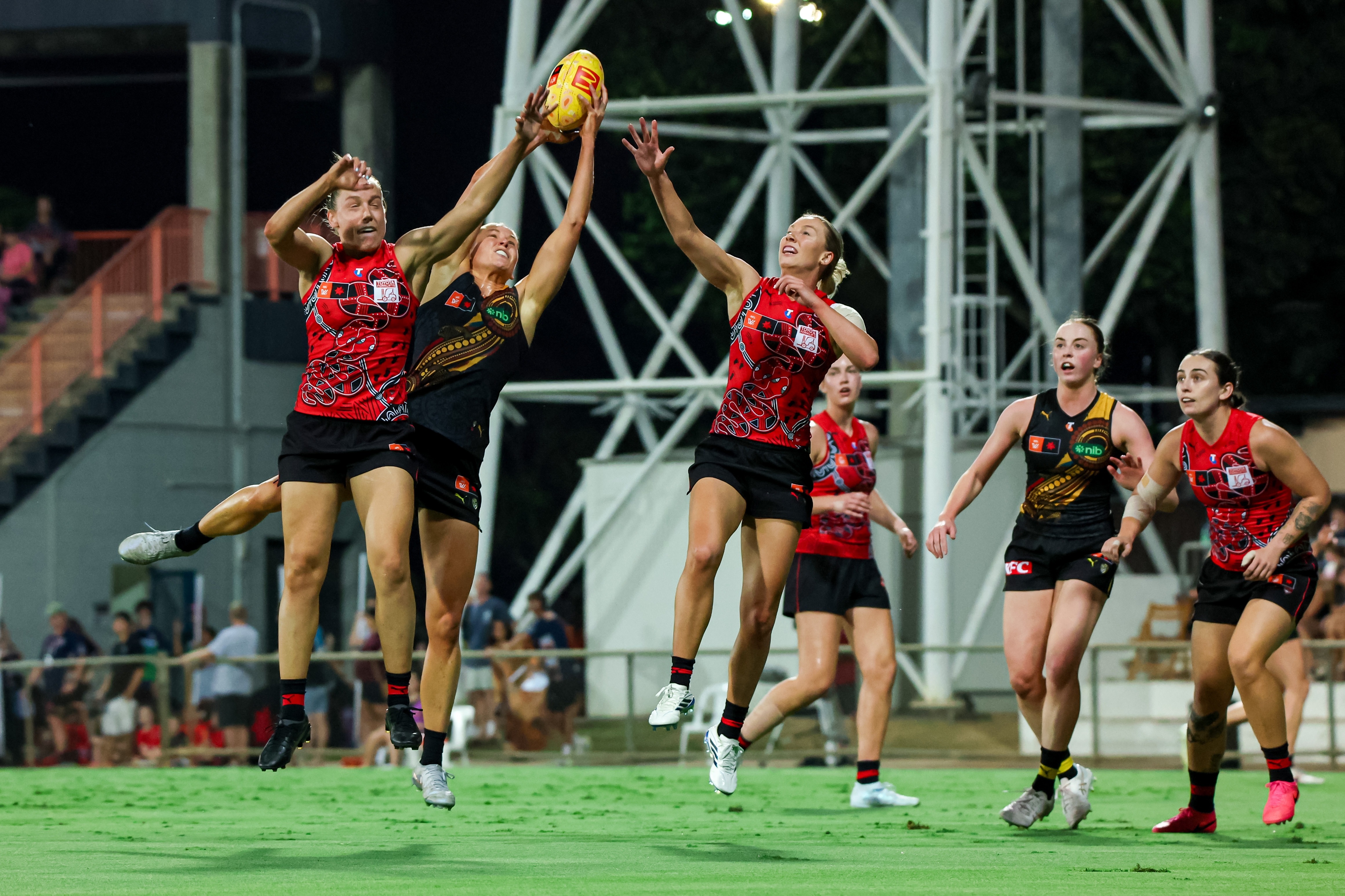 A group of players jump for a yellow AFL ball, two in red and one in yellow and black