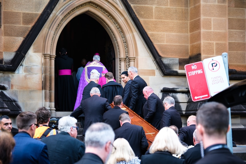 People in black suits carry a casket up stairs toward a wide door