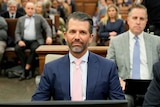 A man in a suit and tie sits at a desk near the front of a courtroom. Other people are behind him in rows of seats.