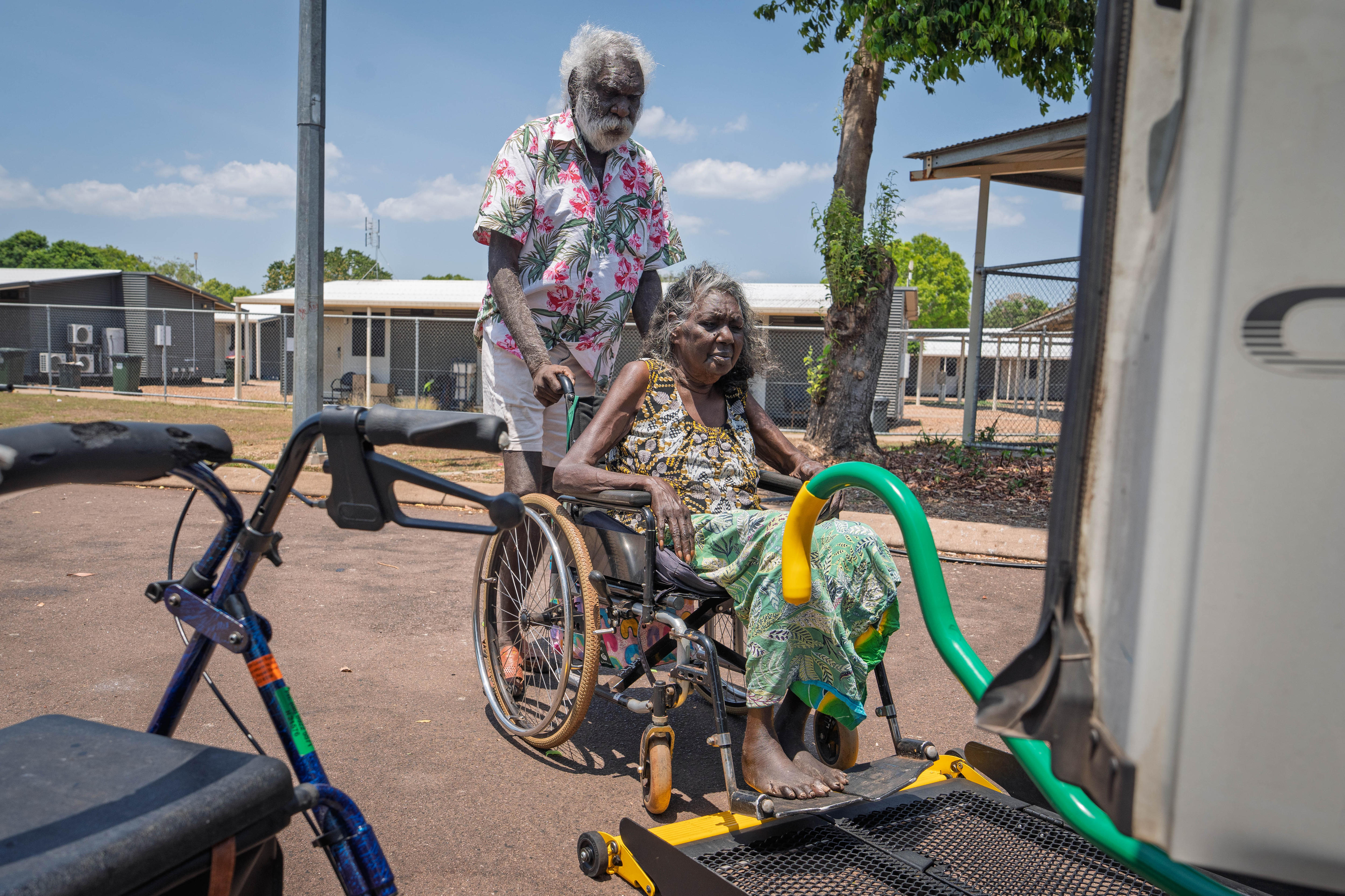 An Indigenous man guides his wife, who is seated in a wheelchair, onto a lift platform attached to the back of a bus.