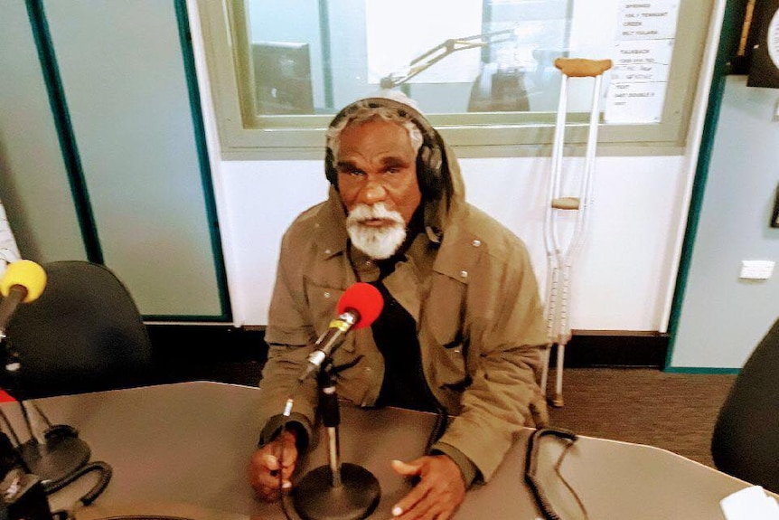 Ned Hargraves sits in a brown jacket talking into a red microphone