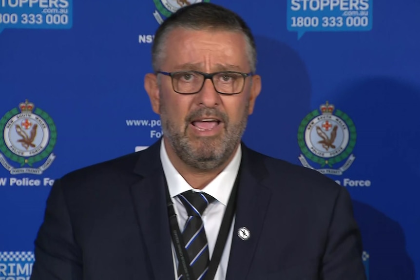 A man wearing a black suit and tie speaks in front of a blue wall with "nsw police force" badges on it