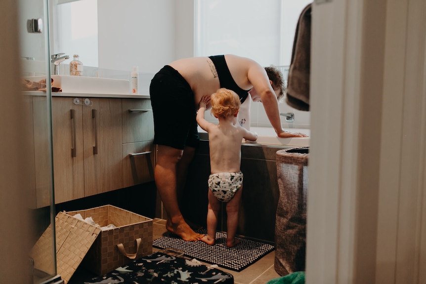 A mother is bent of as she runs a bath while her toddler stands with her.