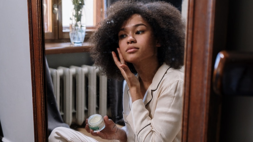 Woman putting face lotion on while looking in mirror for story on for story on skin-whitening cream