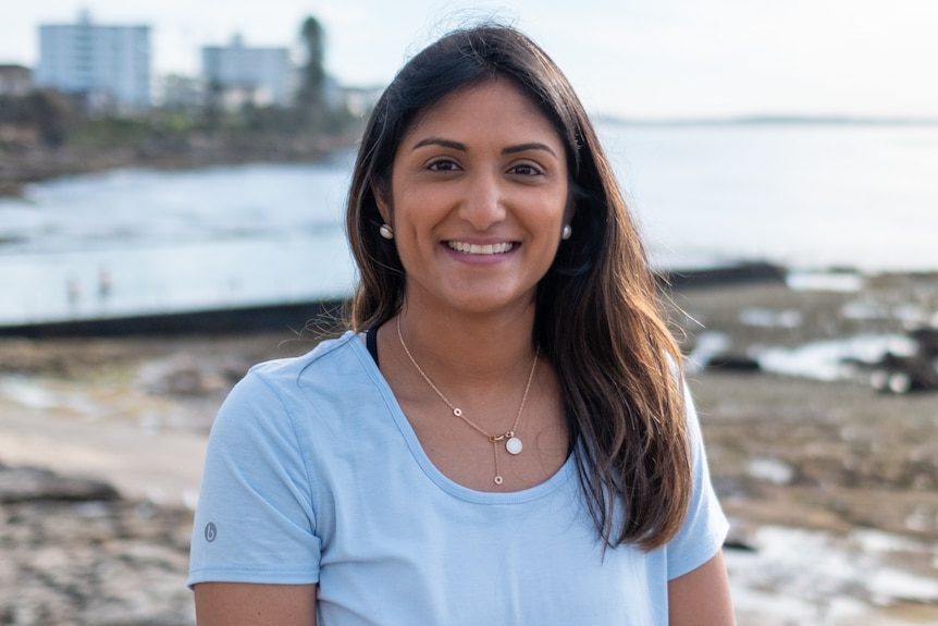 A woman in a blue t-shirt smiles at the camera, the ocean in the background.