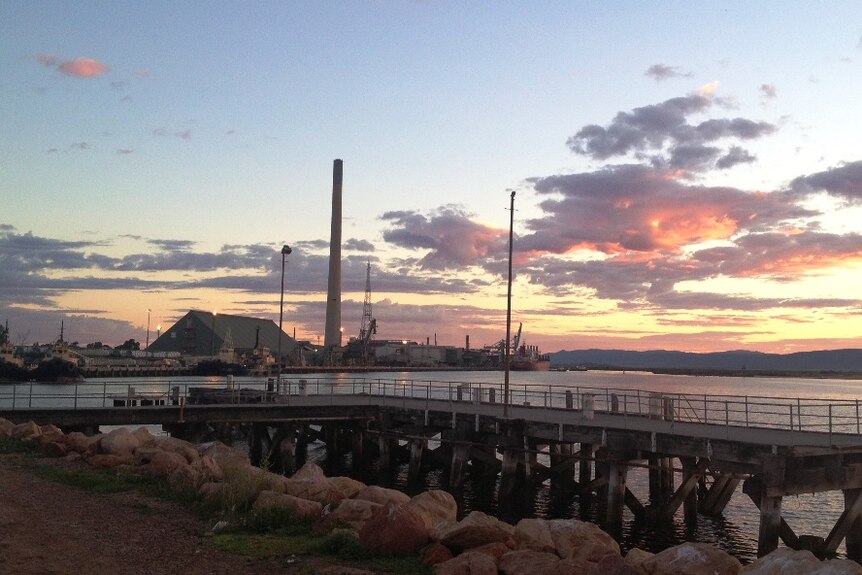 The tall chimney of a large smelter on the water's edge projects into the sky as the sun sets behind it