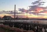 The tall chimney of a large smelter on the water's edge projects into the sky as the sun sets behind it