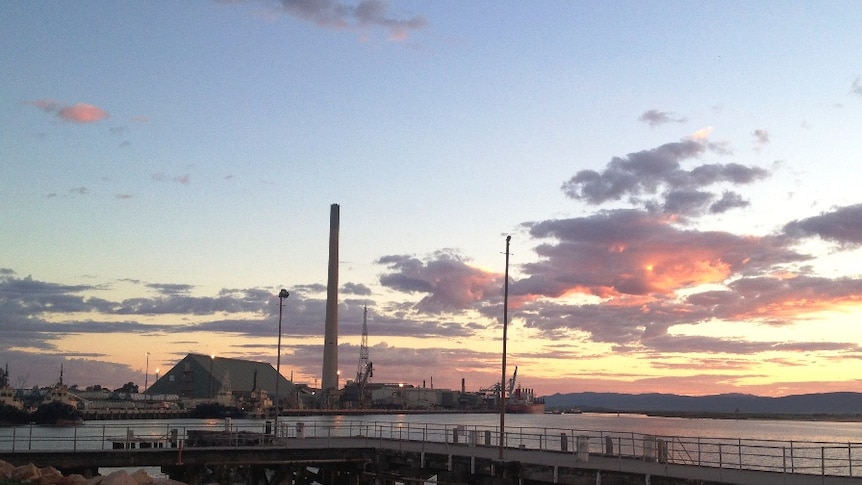 The tall chimney of a large smelter on the water's edge projects into the sky as the sun sets behind it