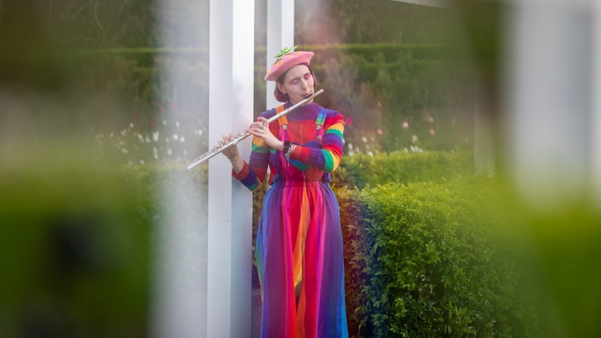 A young woman in pink beret, rainbow jumper and rainbow dress plays a flute in the garden